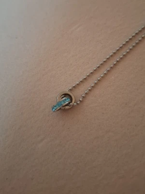 Silverhalsband med blå detalj - Snyggt halsband i silverfärg med kulkedja och ett runt hänge. Hänget har en bågformad detalj med små blå stenar som ger en fräsch touch. Perfekt accessoar för att lyfta din outfit.