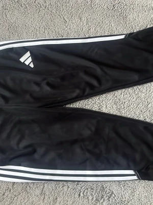 Svarta Adidas träningsbyxor - Säljer ett par svarta träningsbyxor från Adidas med klassiska vita ränder längs sidorna och Adidas-logga på låret. Byxorna har smal passform och är tillverkade i ett lätt och stretchigt material som passar perfekt till träning eller sport.p