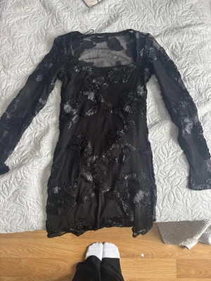 Svart blommig  - Snygg svart kortklänning  i meshmaterial med blommiga broderade detaljer. Klänningen har långärmade transparenta ärmar och en kroppsnära passform. Perfekt för dig som vill sticka ut med coola detaljer och en trendig look.