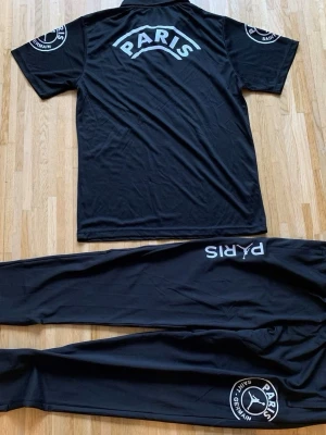 Svart PSG träningsset - Säljer ett svart träningsset från Paris Saint-Germain med tydliga vita loggor och text på både tröja och byxor. Setet består av en kortärmad tröja och matchande byxor i mjukt, sportigt material. Perfekt för fotbollsträning eller chill.