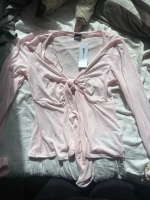 Ljusrosa knytblus från Gina Tricot - Trendig ljusrosa blus från Gina Tricot med långa ärmar och knyt framtill. Tunn och mjuk i materialet, perfekt för lager på lager eller som statement-plagg. Snygg v-ringning och croppad passform som ger en modern vibe.