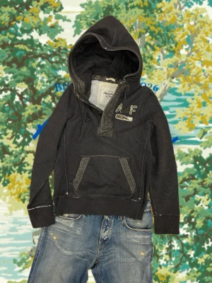 Mörkgrå hoodie från Abercrombie & Fitch - Snygg mörkgrå hoodie från Abercrombie & Fitch med broderad A&F-logga och NY 2004-detalj på bröstet. Hoodien har en stor huva, magficka och ribbade muddar. Perfekt för en avslappnad och trendig stil.