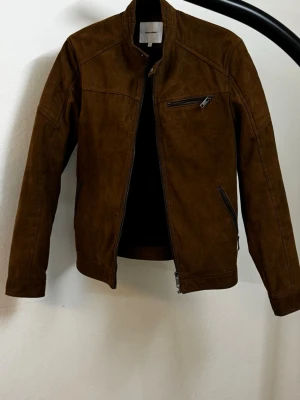 Brun skinnjacka med dragkedjor - Snygg brun skinnjacka från Selected Homme i mocka-look. Jackan har ståkrage, dragkedja framtill och tre fickor med dragkedja. Snygga sömdetaljer och knappdetaljer vid midjan. Perfekt för dig som gillar stilren och tidlös design.