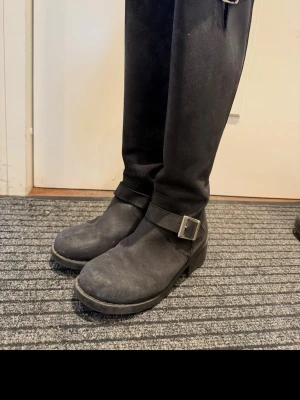 Svarta höga läderstövlar med spänne - Snygga svarta stövlar i läder med högt skaft och grov sula. Stövlarna har dekorativa spännen och dragkedja baktill för enkel på- och avtagning. Perfekta till jeans eller kjol för en cool look.