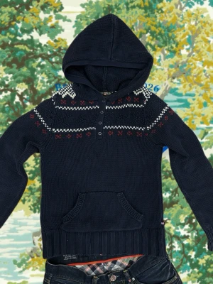Ralph lauren stickad hoodie - Mysig mörkblå stickad hoodie från Ralph lauren med vit och röd jacquardmönstring över axlarna. Tröjan har huva, knappslå vid halsen och en stor magficka. Perfekt för kalla dagar och har en avslappnad passform.