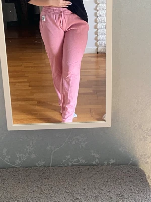 Rosa mjukisbyxor med resårmidja - Säljer ett par ljusrosa mjukisbyxor med resår i midjan och dragsko. Byxorna har raka ben och en liten logga på vänster sida vid höften. Perfekta för chill dagar hemma eller när du vill vara bekväm. Materialet känns mjukt och skönt mot huden. Storlek S