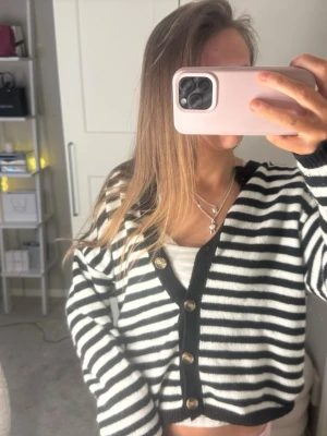 Randig kofta   - Säljer en mysig kofta med svartvita horisontella ränder och svarta kanter. Koftan har v-ringning, guldknappar framtill och är lite croppad i modellen. Perfekt för att lyfta en enkel outfit.