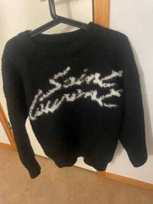 Svart stickad Saint Laurent-tröja - Säljer en svart stickad tröja från Saint Laurent med vit logotyp i graffiti-stil på bröstet. Tröjan har rund halsringning, långa ärmar och ribbade muddar. Perfekt för dig som gillar streetwear och vill ha något riktigt snyggt och bekvämt.