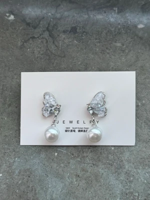 Silverörhängen med fjäril och pärla - Säljer ett par snygga örhängen i silverfärg med glittrande fjärilar upptill och hängande vita pärlor. Fjärilarna har små stenar som ger extra bling och örhängena har stift. Perfekta för dig som gillar söta detaljer och elegant stil.