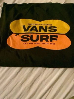 Svart Vans Surf t-shirt med tryck - Cool svart t-shirt från Vans med färgglatt tryck fram och mindre Vans-logga på bröstet. T-shirten har rund hals och är gjord i mjuk bomull. Perfekt för dig som gillar streetwear och surfvibbar.