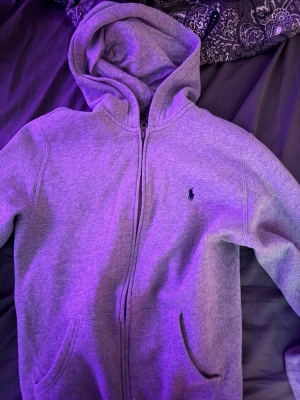 Grå hoodie från Polo Ralph Lauren - Snygg grå zip-hoodie från Polo Ralph Lauren med klassisk huva och dragkedja framtill. Broderad logga i mörkblått på bröstet och två fickor framtill. Perfekt för en chill och stilren look.