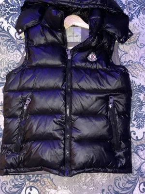 Svart dunväst från Moncler - Snygg svart dunväst från Moncler med glansig finish, två dragkedjeförsedda fickor framtill och ikonisk logotyp på bröstet. Västen har hög krage och quiltad design som ger en modern och sportig look. Perfekt för lager-på-lager-stil.