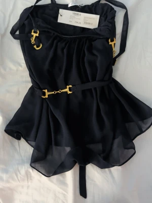 Marinblå chiffontopp Ida Sjöstedt - Säljer en marinblå chiffontopp från Ida Sjöstedt med guldfärgade metalldetaljer och bälte i midjan. Toppen har tunna axelband och ett flowy, elegant fall. Perfekt för dig som gillar unika detaljer och stilren design.