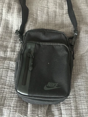 Svart axelväska från Nike - Använd 2 gånger ‼️ Snygg svart axelväska från Nike med justerbar rem och praktiska fack. Väskan har en dragkedja framtill med en diskret Nike-logga och är tillverkad i slitstarkt tyg. Perfekt storlek för mobil, plånbok och andra smågrejer.