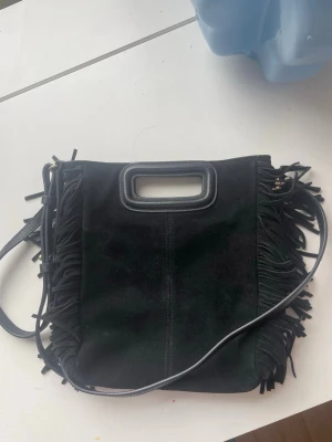 Maje väska i mocka svart - Snygg svart handväska i mocka från Maje med fransar längs kanterna och ett rektangulärt handtag i läder. Väskan har även en axelrem och guldfärgade metalldetaljer. Skriv för mer information 
