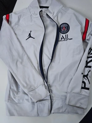 Vit PSG x Jordan ziptröja - Snygg vit ziptröja från PSG x Jordan med svarta och röda detaljer. Tröjan har Paris Saint-Germain-logga på bröstet, Jordan-logga och texten 'PARIS' på ena ärmen. Dragkedja framtill och ribbade muddar. Perfekt för sport eller streetwear.