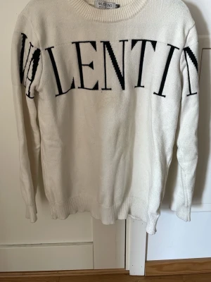 Vit stickad tröja från Valentino - Vit stickad tröja från Valentino med svart, stor logotyp över bröstet och ärmen. Tröjan har rund halsringning och ribbstickade muddar vid ärmslut och nederkant. Tillverkad i Italien och har en klassisk, avslappnad passform.