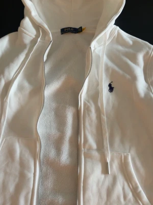 Vit hoodie från Polo Ralph Lauren - Snygg vit hoodie från Polo Ralph Lauren med dragkedja och huva. Klassisk logga i mörkblått broderad på bröstet. Tillverkad i mjukt material med fleecefoder på insidan och praktiska fickor framtill. Perfekt för en chill och stilren look.