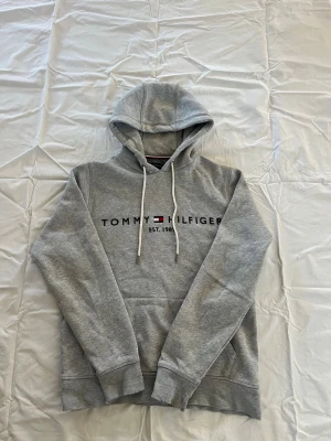 Grå hoodie från Tommy Hilfiger - Snygg grå hoodie från Tommy Hilfiger med klassisk logga och texten 'EST. 1985' på bröstet. Hoodien har huva med vita dragsnören och en stor magficka. Tillverkad i mjukt bomullsmaterial och har ribbade muddar vid ärmslut och nederkant.