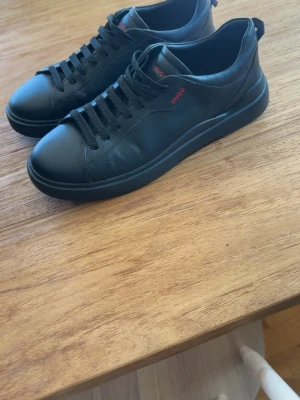 Svarta sneakers från Hugo - Stilrena svarta sneakers från Hugo i slät läderlook med svarta skosnören och diskret röd logga på sidan. Klassisk låg modell med rund tå och svart sula. Perfekta för en clean och modern stil.
