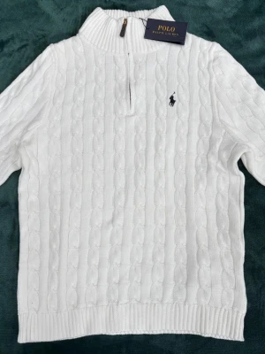 Vit kabelstickad tröja Polo Ralph Lauren - Kabelstickad vit tröja från Polo Ralph Lauren med halv dragkedja och hög krage. Klassisk svart broderad logga på bröstet. Tröjan har ribbade muddar och är perfekt för en clean och stilren look.