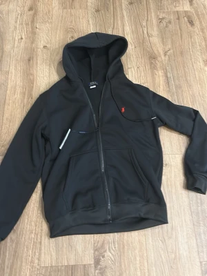 Svart hoodie från Polo Ralph Lauren - Snygg svart hoodie från Polo Ralph Lauren med dragkedja framtill och klassisk röd logga på bröstet. Hoodien har två fickor fram och en skön huva. Perfekt för en avslappnad och stilren look.