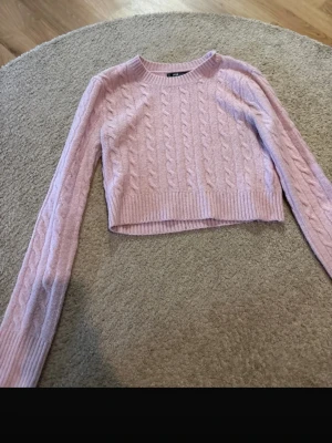 Ljusrosa kabelstickad cropped tröja - Supersöt ljusrosa kabelstickad tröja med croppad passform och långa ärmar. Tröjan har ribbade muddar vid ärmslut och nederkant samt rund halsringning. Perfekt för dig som gillar en mjuk och trendig look.