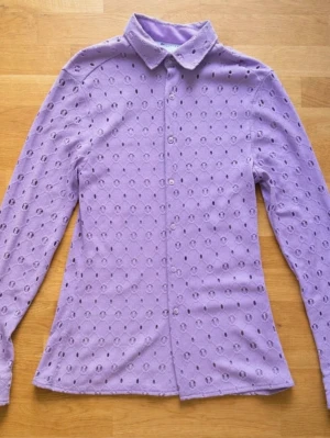Lila skjorta med hålmönster - Säljer en lila skjorta med hålmönster och knappar framtill. Skjortan har klassisk krage och är långärmad. Materialet känns lätt och luftigt, perfekt för dig som vill sticka ut lite extra. Passformen är normal och skjortan har en unik, trendig look. ALDRIG ANVÄND!