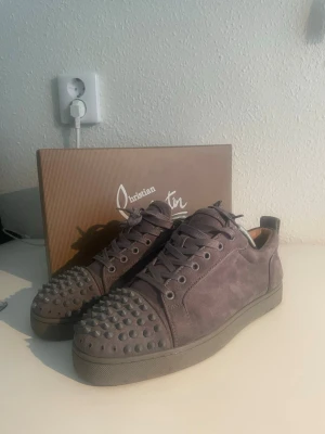 Gråa Christian Louboutin sneakers - Snygga gråa sneakers från Christian Louboutin i mocka med ikoniska nitar på tån. Skorna har lågt skaft, matchande grå snörning och en diskret logga på hälen. Perfekta för dig som vill sticka ut med en edgy och exklusiv stil. OBS! Det saknas några spikar från både höger och vänster sko, men det medföljer att par extra. Annars väldigt fint skick.