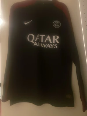 Svart PSG långärmad träningströja Nike - Svart långärmad träningströja från Nike med Paris Saint-Germain-logga på bröstet och Qatar Airways-tryck på magen. Tröjan har röda detaljer längs axlarna och är tillverkad i ett sportigt, lätt material som passar perfekt till fotbollsträning.