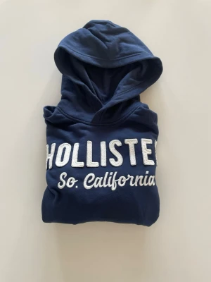 Helt ny, Mörkblå hoodie från Hollister - HELT NY med prislapp, Snygg mörkblå hoodie från Hollister med stor vit broderad text 'HOLLISTER So. California' på bröstet. Klassisk huva och mjukt material som känns skönt mot huden. 