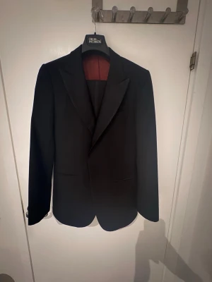 Svart smoking från Suitsupply, storlek 46, allt inkluderar. - Klassisk svart kavaj från Suitsupply i 100% ull med elegant passform och slät design. Fodret är vinrött och kavajen har spetsiga slag samt diskreta fickor. Perfekt för dig som vill ha en stilren och tidlös look.