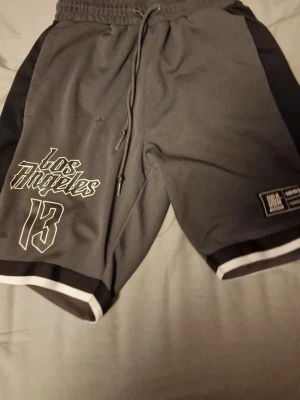 Grå Los Angeles basketshorts - Säljer ett par grå basketshorts från UGA med svart och vit kant nertill, snörning i midjan och tryck med 'Los Angeles' och siffran 13 på ena benet. Shortsen har elastisk midja och är perfekta för sportiga outfits.