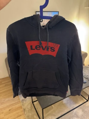 Mörkblå hoodie från Levi's med logga - Cool blå hoodie från Levi's med stor röd logga på bröstet. Klassisk modell med huva, snören och en stor magficka. Perfekt för dig som gillar streetwear och vill ha något bekvämt och snyggt. Materialet är mjukt och känns skönt mot huden.