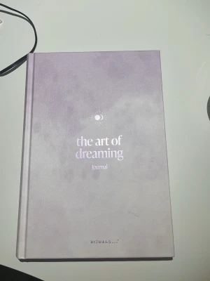 The Art of Dreaming Journal - En snygg drömdagbok med hård pärm i ljuslila nyans från Rituals. Boken har inspirerande citat och instruktioner för att skriva ner och utforska sina drömmar. Inlagan har färgglada illustrationer med moln och fåglar, vilket ger en kreativ och drömsk känsla. Aldrig använd eller läst men den är år gammal