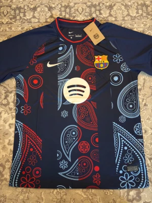 Barcelona Nike matchtröja paisley - Snygg Barcelona matchtröja från Nike med paisleymönster i rött och ljusblått på mörkblå botten. Tröjan har klubbmärke, Nike-logga och Spotify-logga på bröstet. Tillverkad i lätt och funktionellt material, perfekt för fotboll eller träning.