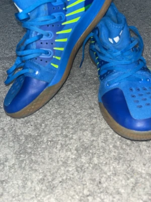 Blå inomhusskor från Adidas - Säljer ett par blå inomhusskor från Adidas med coola neongröna detaljer och gummisula. Skorna har snörning, ventilerande hål på ovandelen och en slitstark sula som passar perfekt för sporter inomhus. Adidas-logga syns på plösen.