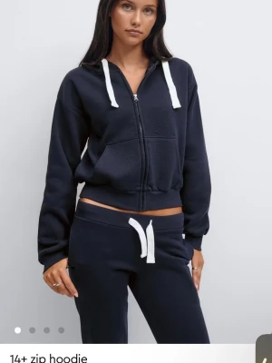 Mörkblå zip hoodie med vita snören - Snygg mörkblå hoodie med dragkedja och vita snören i huvan. Hoodien har en croppad passform, två stora fickor framtill och ribbade muddar vid ärmar och nederkant. Perfekt för en avslappnad och trendig look.