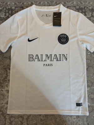 Vit PSG x Balmain tröja Nike - Snygg vit t-shirt från Nike i samarbete med PSG och Balmain. Tröjan har svart Nike-logga, PSG-emblem och Balmain Paris-tryck på bröstet. Tillverkad i lätt och ventilerande material, perfekt för träning eller streetwear.