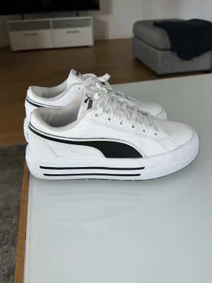 Vita Puma sneakers med svart detalj - Snygga vita sneakers från Puma med svart logga på sidan och svarta ränder på den tjocka sulan. Skorna har lågt skaft, vita snören och är tillverkade i textil och syntet. Perfekta för dig som gillar klassisk streetstyle.