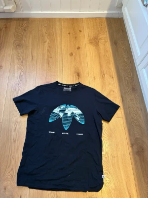 Adidas t-shirt  - Snygg marinblå t-shirt från Adidas med det klassiska treklöver-trycket i blått och vitt framtill. Modellen har rund hals och korta ärmar. Tillverkad i mjuk bomull som känns skön mot huden. Perfekt för dig som gillar streetwear och sportig stil.