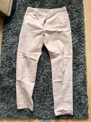 Ljusrosa chinos med smal passform - Säljer ett par ljusrosa chinos med smal passform och klassiska fickor fram. Byxorna har bälteshällor och är tillverkade i ett mjukt bomullsmaterial som känns skönt mot huden. Perfekta för dig som vill ha en clean och stilren look.