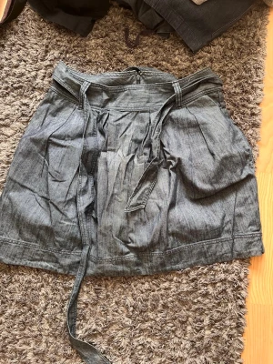 Blå kortkjol med knytband - Säljer en blå kortkjol i denim-look med knytband i midjan. Kjolen har veckade detaljer framtill och dragkedja bak. Perfekt för dig som gillar en avslappnad men ändå snygg stil.