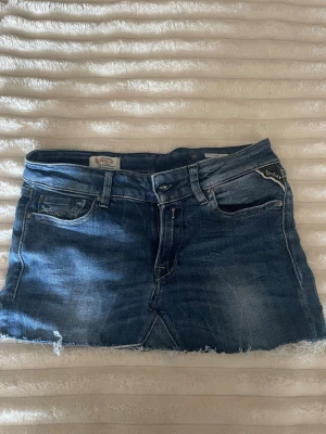Blå jeanskjol från Replay - Säljer en snygg blå jeanskjol från Replay med klassisk femficksdesign och rå kant nertill. Skorten har en cool tvättad look och detaljer som Replay-logga på fickan. Perfekt för dig som gillar avslappnad denimstil.