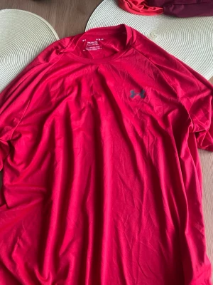 Röd t-shirt från Under Armour - Snygg röd t-shirt från Under Armour med klassisk logga på bröstet. Tillverkad i ett lätt och mjukt material som andas, perfekt för träning eller chill. T-shirten har korta ärmar och en relaxed fit som ger skön rörelsefrihet.