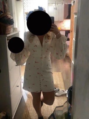 Vit kortklänning med blommor adoore - Söt vit kortklänning med små röda blommor, v-ringning och knappar framtill. Klänningen har långa puffärmar med resår vid handlederna och en figurnära passform. Perfekt för sommarens alla tillfällen.