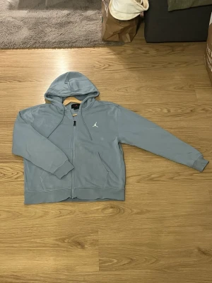 Ljusblå hoodie från Jordan - Ljusblå hoodie med dragkedja från Jordan. Klassisk design med huva och dragsnören, två fickor framtill och ribbade muddar. Tillverkad i mjukt bomullsmaterial och har den ikoniska Jumpman-loggan broderad på bröstet.