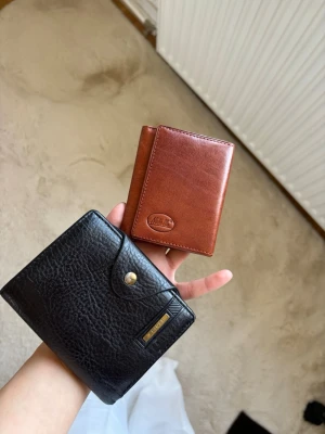 Två stilrena läderplånböcker - Säljer två plånböcker i äkta läder. En är svart med knappstängning och metallogo från Wild Tiger, och den andra är brun med enkel design och präglad logga. Båda har flera fack för kort och sedlar, perfekta för dig som vill ha koll på dina grejer.