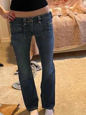 Mörkblå lågmidjade  bootcut jeans med detaljer - Säljer ett par mörkblå bootcut jeans med snygga slitningar och kontrastsömmar. Jeansen har fem fickor, dekorativa nitar och en liten logga på bakfickan. Klassisk knappgylf och cool tvätt som ger en avslappnad vibe.