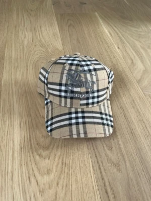 Beige rutig keps från Burberry - Säljer en klassisk beige keps från Burberry med det ikoniska rutmönstret i svart, vitt och rött. Kepsen har böjd skärm och broderad logga framtill med en riddare. Justerbar baktill för perfekt passform. Perfekt accessoar för en trendig och stilren look.